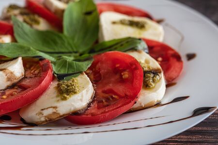 White plate of healthy classic delicious caprese salad with tomatoes and mozzarella cheese with basil leaves and strips of soy sauce on the wood background. Close up.の写真素材