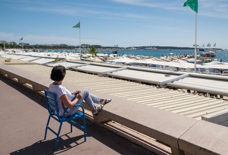 European woman sitting in a chair on the waterfront of Cannes and looking at the sea on a clear sunny dayの写真素材
