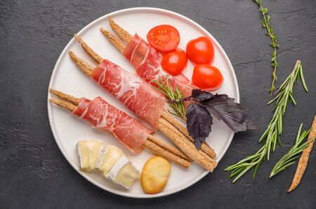 Traditional Italian food is grissini bread with prosciutto, cheese and tomatoes with herbs on a plate on a dark background. Healthy style. Copy spaceの写真素材