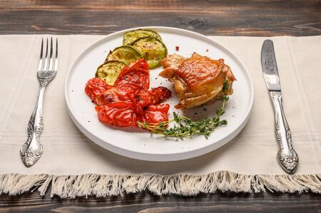 Homemade baked chicken thigh, zucchini and peppers with soy sauce, spices and herbs on a white plate on a linen napkin with a knife and fork. Rustic styleの写真素材