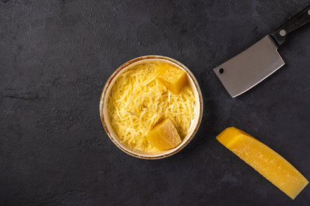 Grated parmesan in ceramic bowl, pieces of cheese and knife on a dark graphite background, top view,...