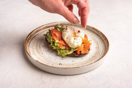 Womans hand puts pepper on poached egg toast with avocado, salted salmon, arugula and rye bread on a ceramic plate, close up. Healthy food conceptの写真素材