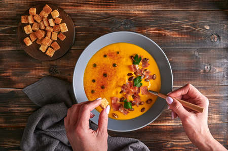 Womans hands hold crouton and spoon with traditional pumpkin puree cream soup with carrots, onions, ginger, garlic, coconut milk. Added seeds, fried bacon, parsley. Healthy, vegetarian food. Top viewの写真素材