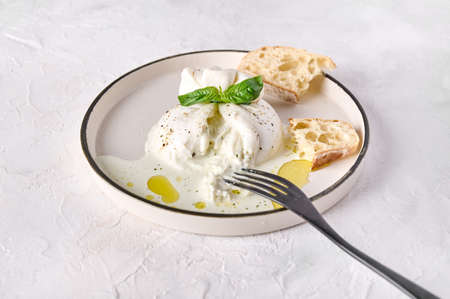 Italian cut burrata cheese with ciabatta bread and olive oil on white plate with black fork. Close...