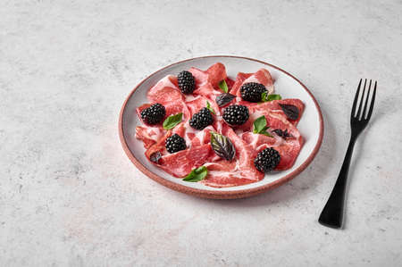 Close up slices of prosciutto ham coppa di parma with basil and blackberry on light plate and black fork with copy spaceの写真素材