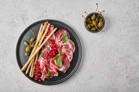 Slices of prosciutto coppa di parma ham with basil, grissini sticks, capers and redcurrant on dark plate on textured background. Top view with copy spaceの写真素材