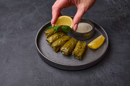 Womans hand squeezes lemon on dolma with parsley, saucepan with sour cream and lemon in black plate on dark wood background. Close up. Selective focusの写真素材