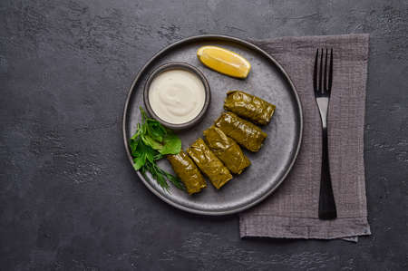 Flat lay dolma, parsley, saucepan with sour cream and lemon in a black plate on a dark table, near black fork and linen napkinの写真素材