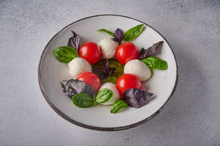 Homemade caprese salad with ripe tomatoes, fresh basil and mozzarella cheese in white plate on grey...