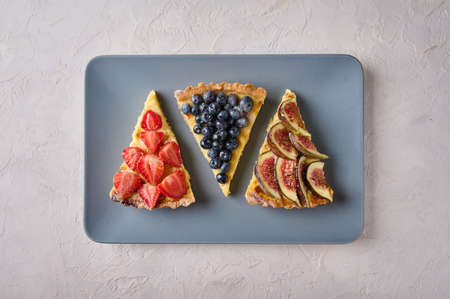 Three desserts of tart cake with strawberries, blueberries and figs on rectangular plate on light textured background. Top viewの写真素材