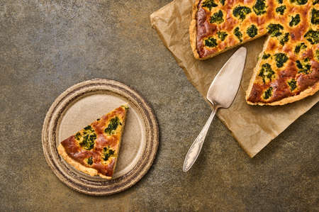Piece of homemade quiche on ceramic rustic plate. Nearby is large pie on parchment paper with...