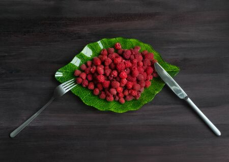 Ripe raspberry in a plate in the form of a green leaf on a black wooden background with a fork and a knife. Fresh berries. Concept Useful vitamin fruit diet. Fresh natural Breakfast. Vegan. copyspaceの写真素材