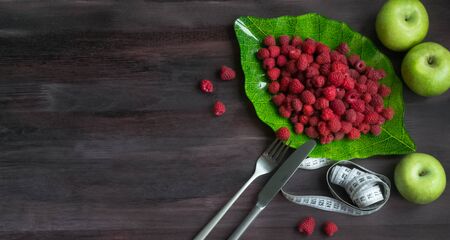 Ripe raspberry in a green leaf plate on a black wooden background with fork, knife, apples and measure tape. Fresh berries. Concept Useful vitamin fruit diet. Natural Breakfast. Vegan. copyspaceの写真素材