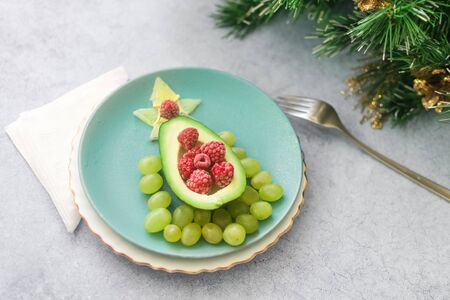 Delicious edible Christmas tree of avocado, frozen sweet raspberries and grapes on a blue plate on...