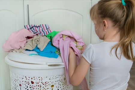 Dirty baby clothes.A small blonde girl in a white t-shirt puts clothes in a Laundry basket.Washing and cleaning.Homework.House cleaningの写真素材