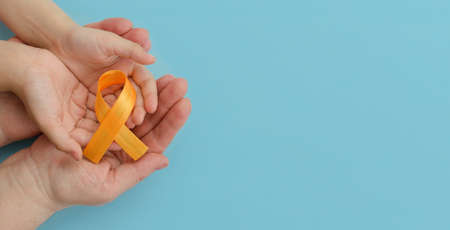 Hands of mother and child hold orange ribbon on blue background.Concept. World Multiple Sclerosis day.Layout. Copy space. Banner. Mock upの写真素材