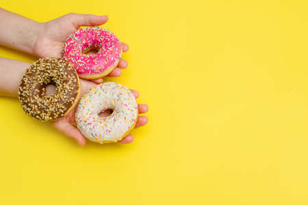 Hands hold delicious sweet donuts with frosting and filling on a yellow background. Favorite American pastry. A greeting card or banner for sale on a website or in a store.Copy spaceの写真素材
