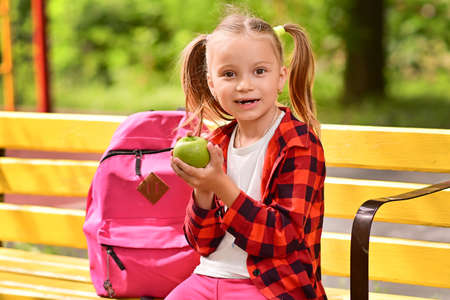 Back to school. Cute little schoolgirl is sitting on bench in school yard and biting green apple. Child with backpack is eating from lunch box outdoor. Right school meal for lunch. Students new yearの写真素材