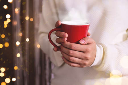 Merry Christmas. Woman in white sweater hands holding christmas hot drink or cofee in red cup. Shimmer background with snow and light bokeh. Greating card or bannerの写真素材