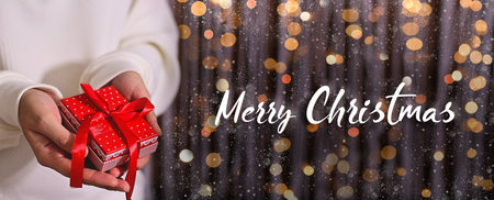Merry Christmas. Woman in white sweater hands holding red gift box on shimmer background with snow and light bokeh. Greating card or banner with text.の写真素材