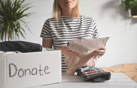 Young woman stands at a table in room and puts clothes in a donation box. Clothes Donation, Renewable Concept and reuse recycleの写真素材