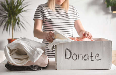 Young woman stands at a table in room and puts clothes in a donation box. Clothes Donation,...