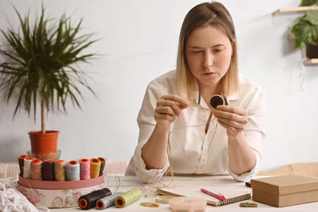 A young blonde woman stands at a table and does macrame needlework, creates home decorations. Small business. Master of jewelry weaving.の写真素材