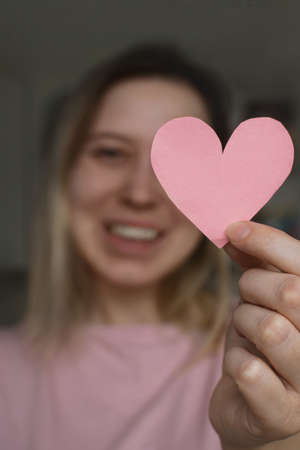 Young woman in pink T-shirt holds pink heart in her hand at level of her face and smiles. Gift...