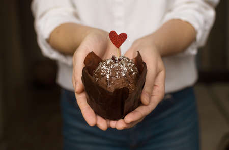 Young woman in white shirt holds a delicious chocolate cupcake with sprinkles and red heart. Authentic dark photography. Gift greeting for holiday. Valentine's Day, Mother's Day, Women's Day.の写真素材