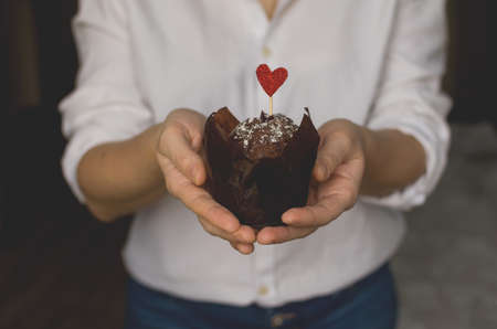 Young woman in white shirt holds a delicious chocolate cupcake with sprinkles and red heart. Authentic dark photography. Gift greeting for holiday. Valentines Day, Mothers Day, Womens Dayの写真素材
