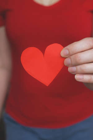 soft focus. Young woman in a red T-shirt holds a red heart cut out of paper. Authentic photo. close-up. Valentins Day, Mother's Day, March 8 or Women's Day. greeting card. verticallyの写真素材