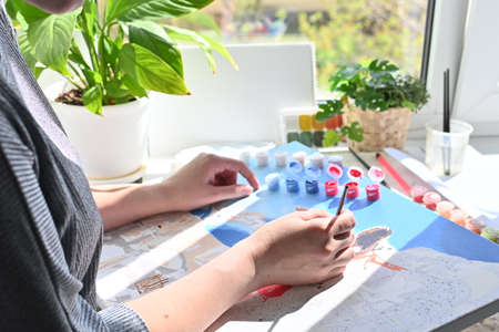 Leisure time. Painting a picture of a house with paints on canvas. A young woman paints a picture near the window with paints. Home hobby, recreationの写真素材