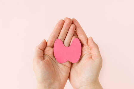 World Thyroid Day. Womens hands hold paper form of the thyroid gland on pink background. Problems...