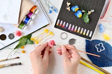 Leisure. Hobbies and recreation. Creating flowers and ornaments from cold porcelain. Painting of jewelry. Womans hands hold flower made of cold porcelain and brush against background of materialsの写真素材