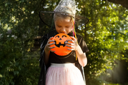 Happy Halloween day. Little girl dressed in a witch costume holds bucket of sweets and looks at the...