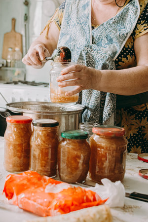 An elderly woman puts jam in a jar for preservation. Red ripe apples on the table next to the jam pot. An elderly woman makes jam in her cottage on the farm.の写真素材