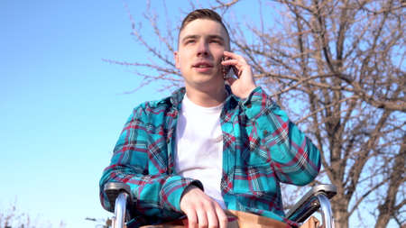 A young man on a wheelchair is talking on the phone. Disabled man in nature with a smartphone.