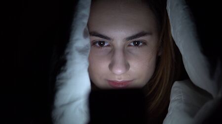 A young woman lies under a blanket with a phone in her hands. A woman in the dark uses a pad. Modern...
