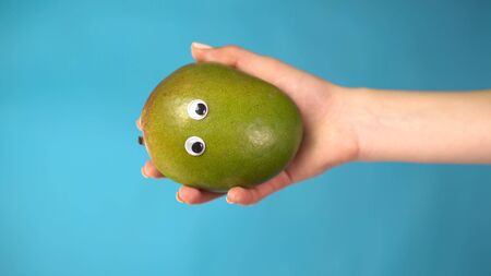 Mango with eyes in a woman hand on a blue background. Woman hand close-up. Vertical photo.の写真素材