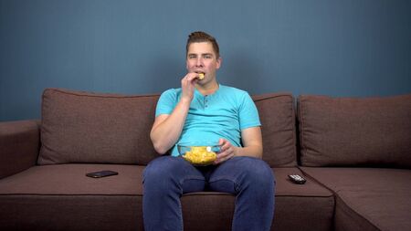 A young man sits on a sofa eating chips and watching TV. The view from the TV. Looking at the cameraの写真素材