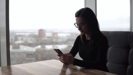 A young woman with a phone sits at a table. A girl sits in a cafe by the panoramic window with a smartphone in her hands. View from the window.の写真素材