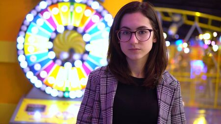 A young woman is standing in an amusement park. The camera moves closer to the girl. Park for children with slot machines and bright light bulbs.の写真素材