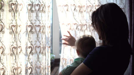 A young mother holds a baby in her arms. A woman with a child looking out the window pushing the curtain.の写真素材