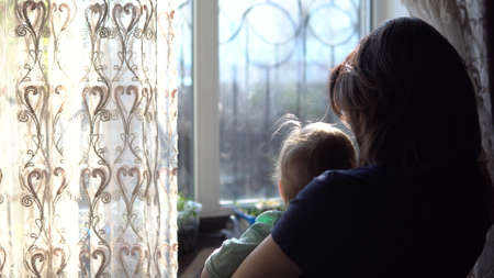 A young mother holds a baby in her arms. A woman with a child looking out the window pushing the curtain.の写真素材