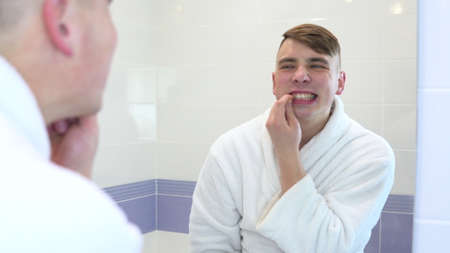 A young man in a white coat looks in the mirror at his teeth. View through the mirrorの写真素材
