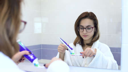 A woman in a white coat smears toothpaste on a brush. Look through the mirror