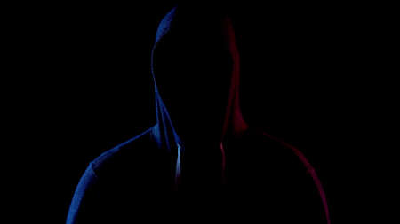 A young man is standing in the hood hiding his face. An attacker is standing in the dark. Blue and red light falls on the person on a black background.の写真素材
