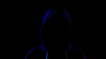 A young woman is standing in the hood hiding her face. An attacker is standing in the dark. Blue and red light falls on the person on a black background.の写真素材