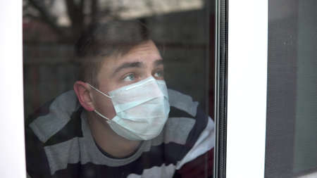 A sad young man is sitting at home in quarantine. A man looks out the window and puts his hand to...