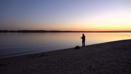 A man is fishing at sunrise by the river. The yellow sun rises from the horizon. Silhouette of a fisherman.の写真素材